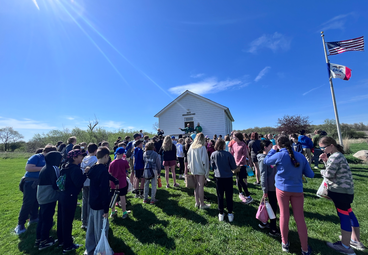 5th graders stand on a sunny lawn outside the borlaug schoolhouse under american and iowa flags