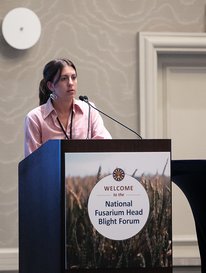 Rae page speaks at a podium with a sign reading "Welcome to the National Fusarium Head Blight Forum"