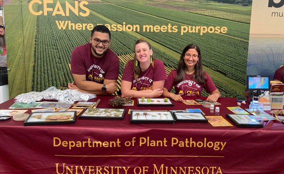 José, Becca, and Sita stand at a maroon table covered in plant disease samples. A field print backdrop reads "CFANS: Where passion meets purpose"