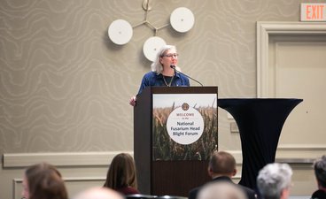 Ruth Dill-Macky speaks to a crowd while standing at a podium labeled Welcome to the National Fusarium Head Blight Forum