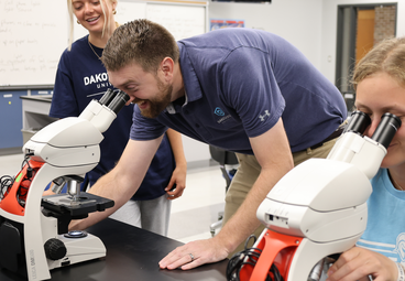 Adnrew sathoff bends over a microscope smiling and talking to two students